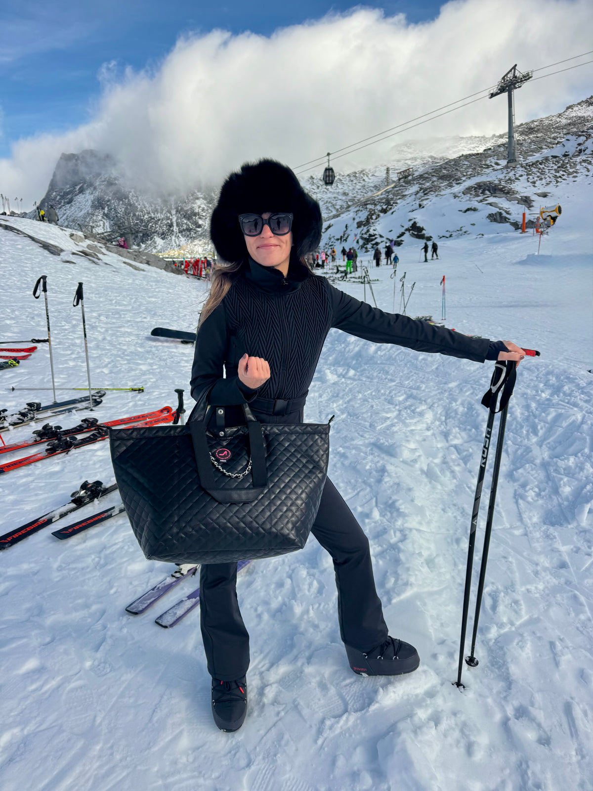 Woman in black ski outfit holds black caviar leather medium tote bag on snowy slope with ski gear.