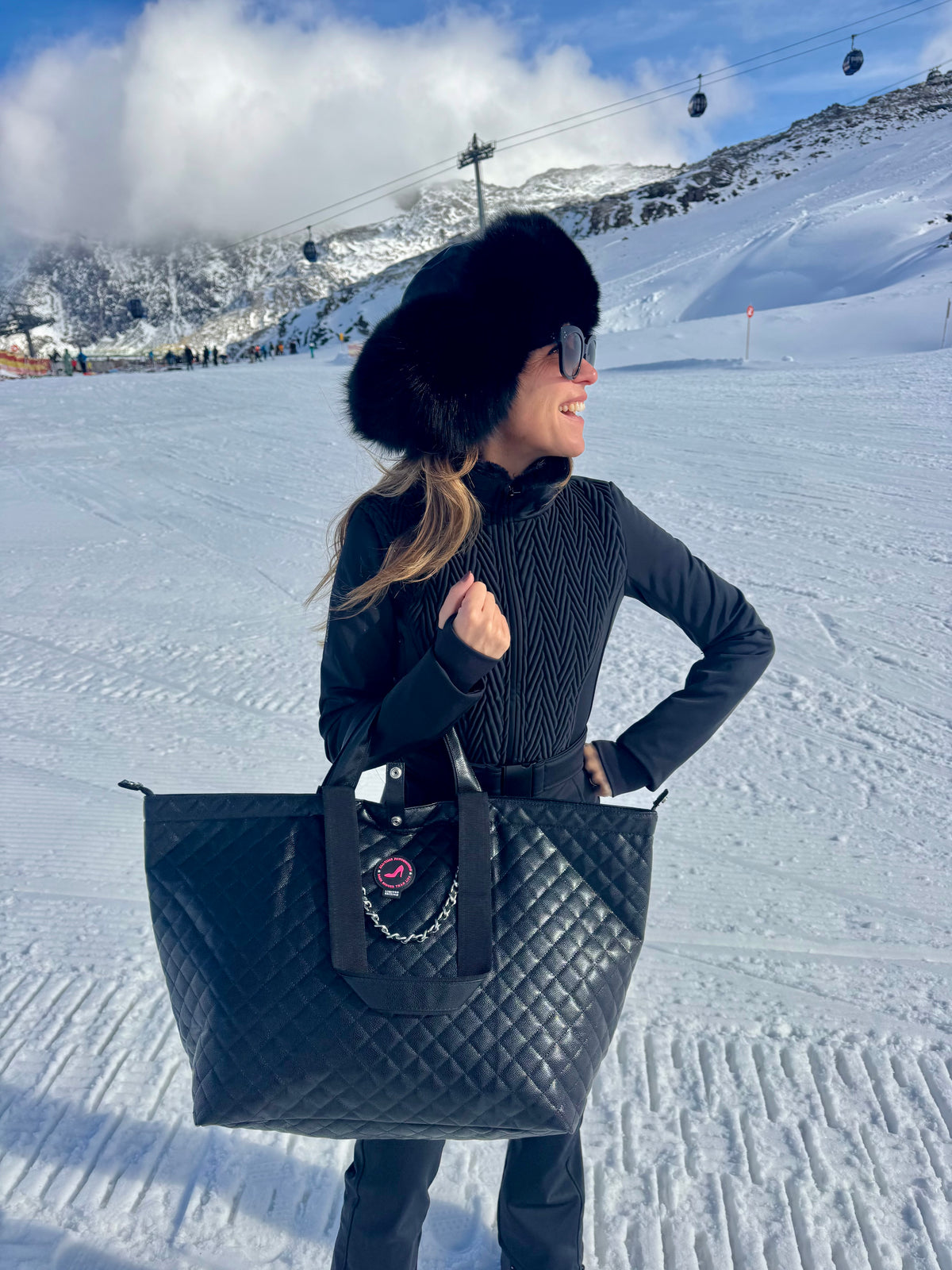 Woman in winter wear holding black caviar leather medium tote bag on snowy mountain with ski lifts in background.