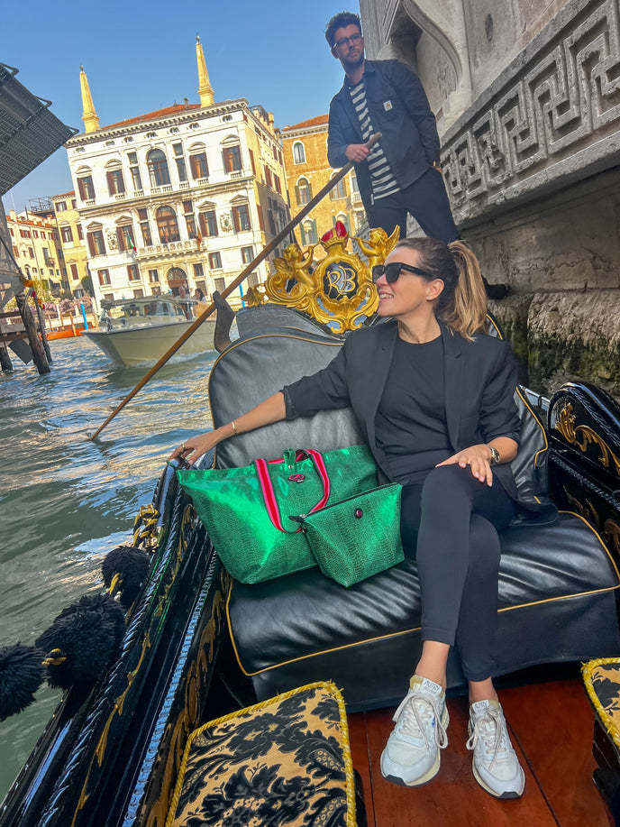 Woman sitting on a gondola in Venice with the Medium Tote Bag Venezia in metallic green croc print and matching Toiletry Bag, featuring pink and red striped handles and signature logo detail.