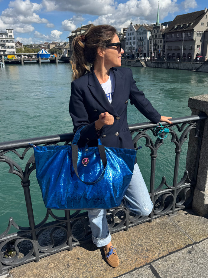 Woman with a blue bag standing by a waterfront with buildings in the background