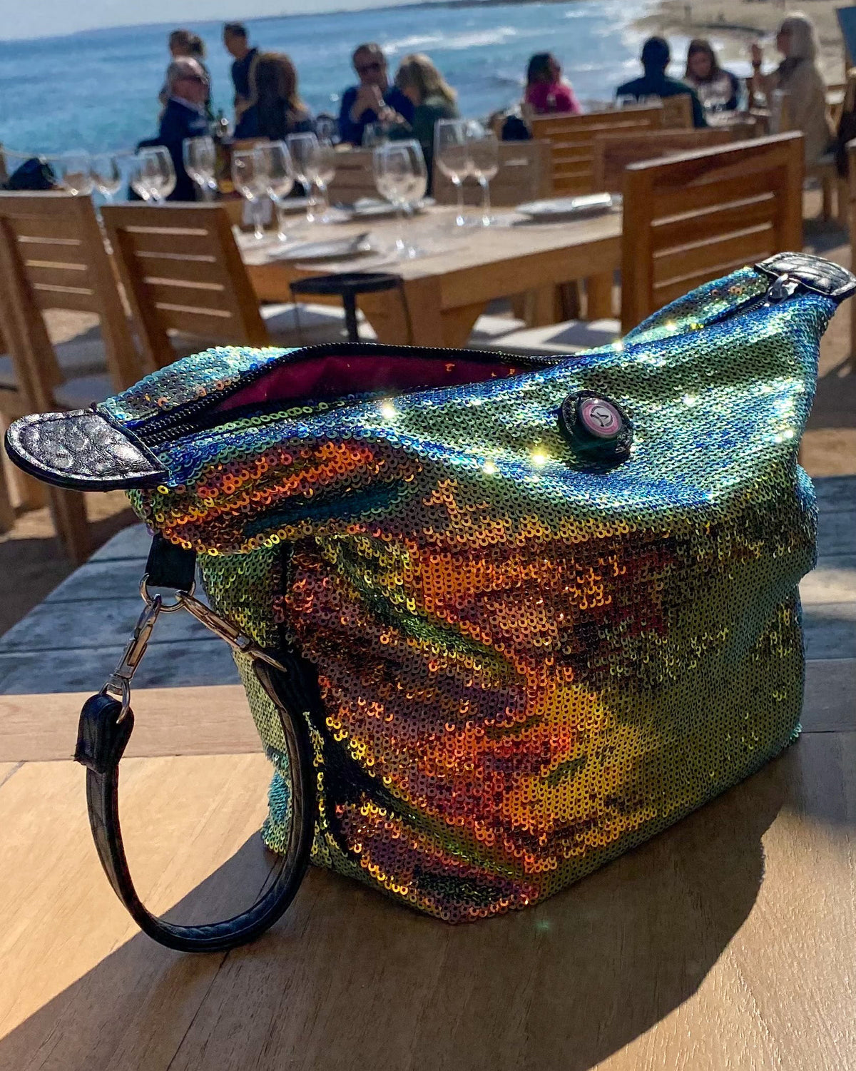 A large, iridescent sequined clutch with a black leather strap sits on a wooden table at an outdoor beachside restaurant, with the ocean and dining area blurred in the background.