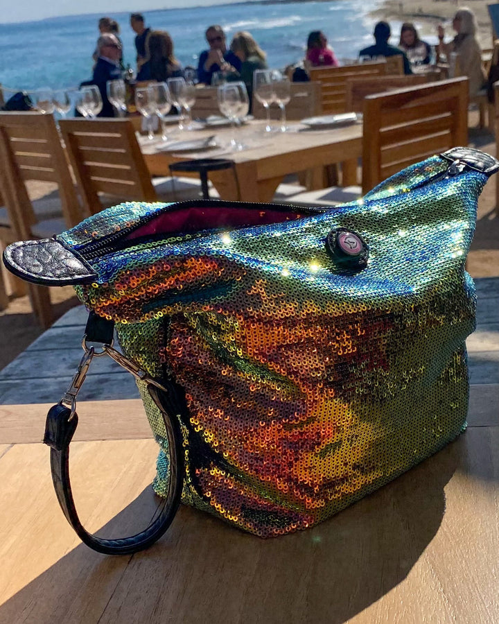 A large, iridescent sequined clutch with a black leather strap sits on a wooden table at an outdoor beachside restaurant, with the ocean and dining area blurred in the background.
