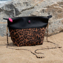 Leopard print handbag with a black top section on a sandy surface with rocks and water in the background