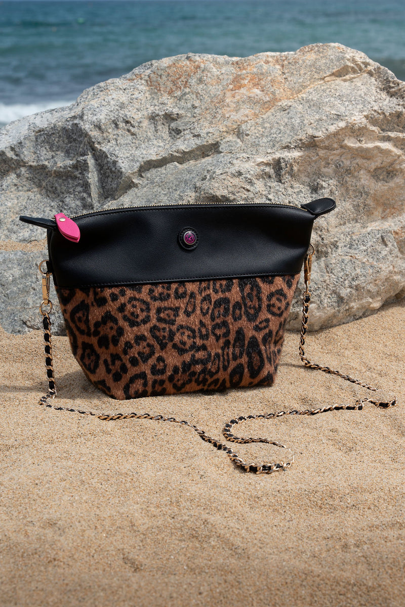 Leopard print handbag with a black top section on a sandy surface with rocks and water in the background