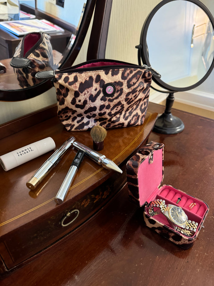 Kosmetiktasche mit Leopardenmuster, darin Pinsel, Lipgloss, ein Stift und eine Schmuckschatulle auf einem hölzernen Schminktisch mit Spiegeln.