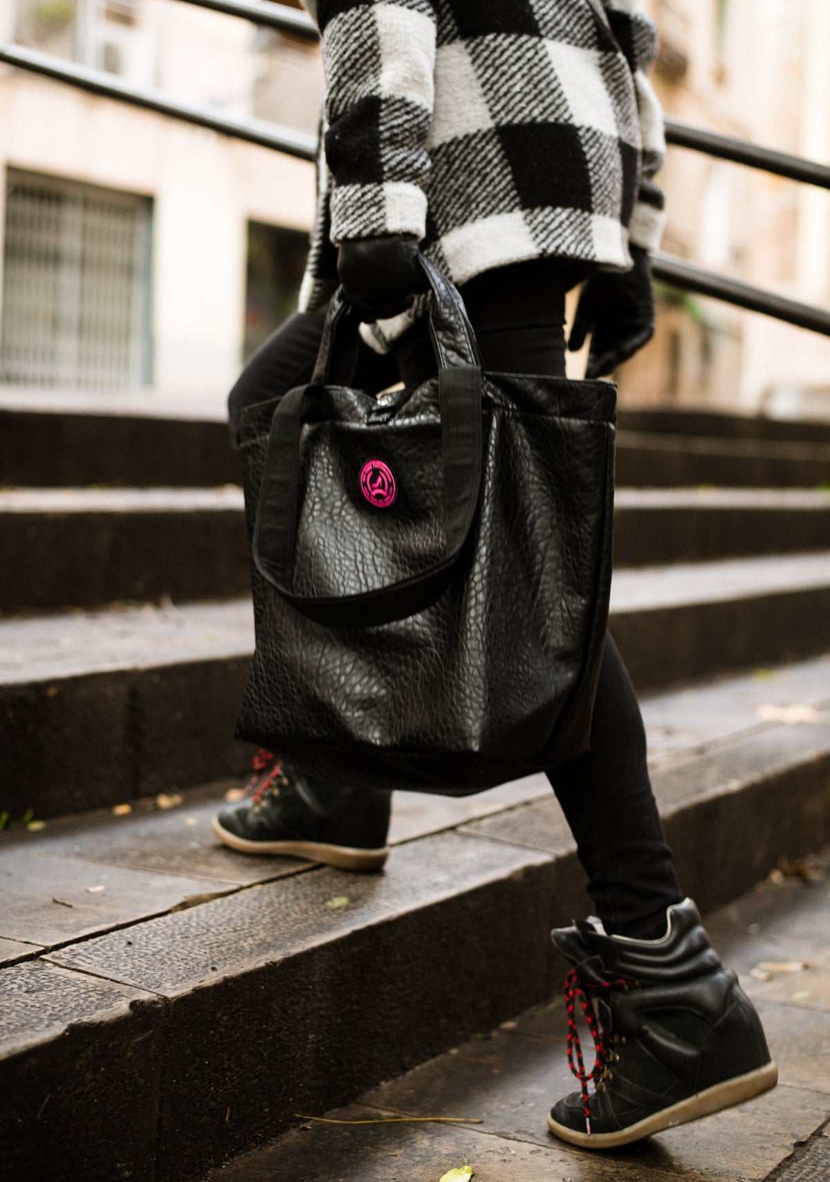 Woman in boots and leggings climbs stairs carrying black croc-embossed leather small tote bag and wearing plaid jacket.