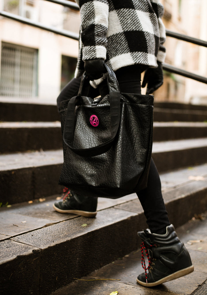 Eine Frau in Stiefeln und Leggings steigt die Treppe hinauf; sie trägt eine kleine schwarze Handtasche aus Leder mit Krokodilprägung und eine karierte Jacke.