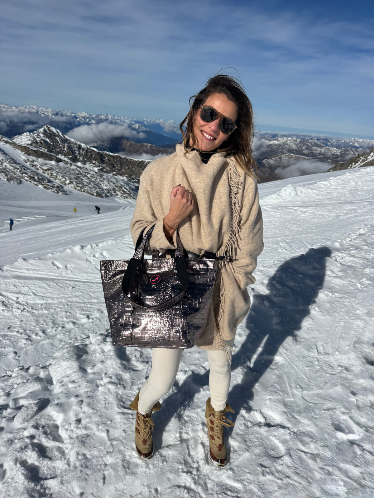 Eine Frau auf einem schneebedeckten Gipfel hält eine kleine BRIGITTE-Tasche in Bronze-Metallic in der Hand, trägt eine Sonnenbrille und ein beiges Outfit.