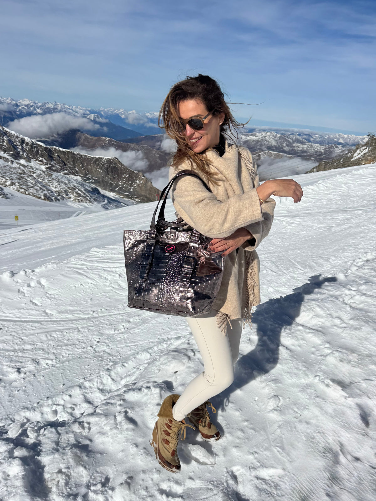 Frau in beiger Winterkleidung mit einer kleinen, bronzefarbenen Metallic-Tragetasche der Marke „All-time Favourites BRIGITTE“ auf einem verschneiten Berg.