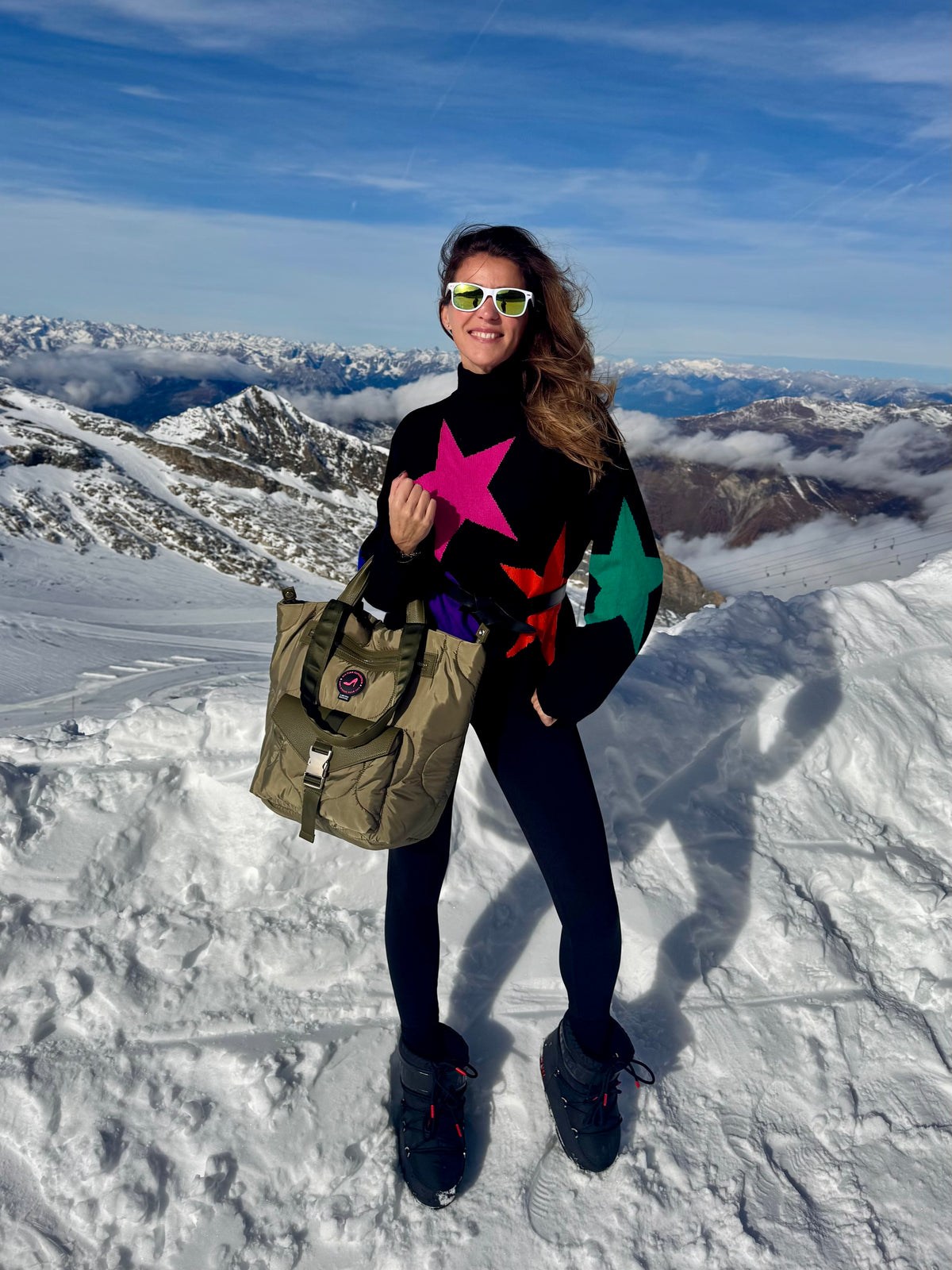 Woman in star sweater holding DIANA All-time Favourites small green puffy tote bag on snowy mountain.