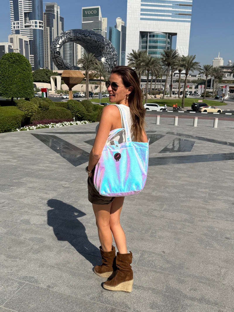 Woman walking in a Dubai city square holding a white pearl–coloured sequin tote bag, with modern buildings and palm trees in the background