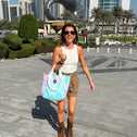 Woman walking in a Dubai city square holding an iridescent tote bag, with modern buildings and palm trees in the background