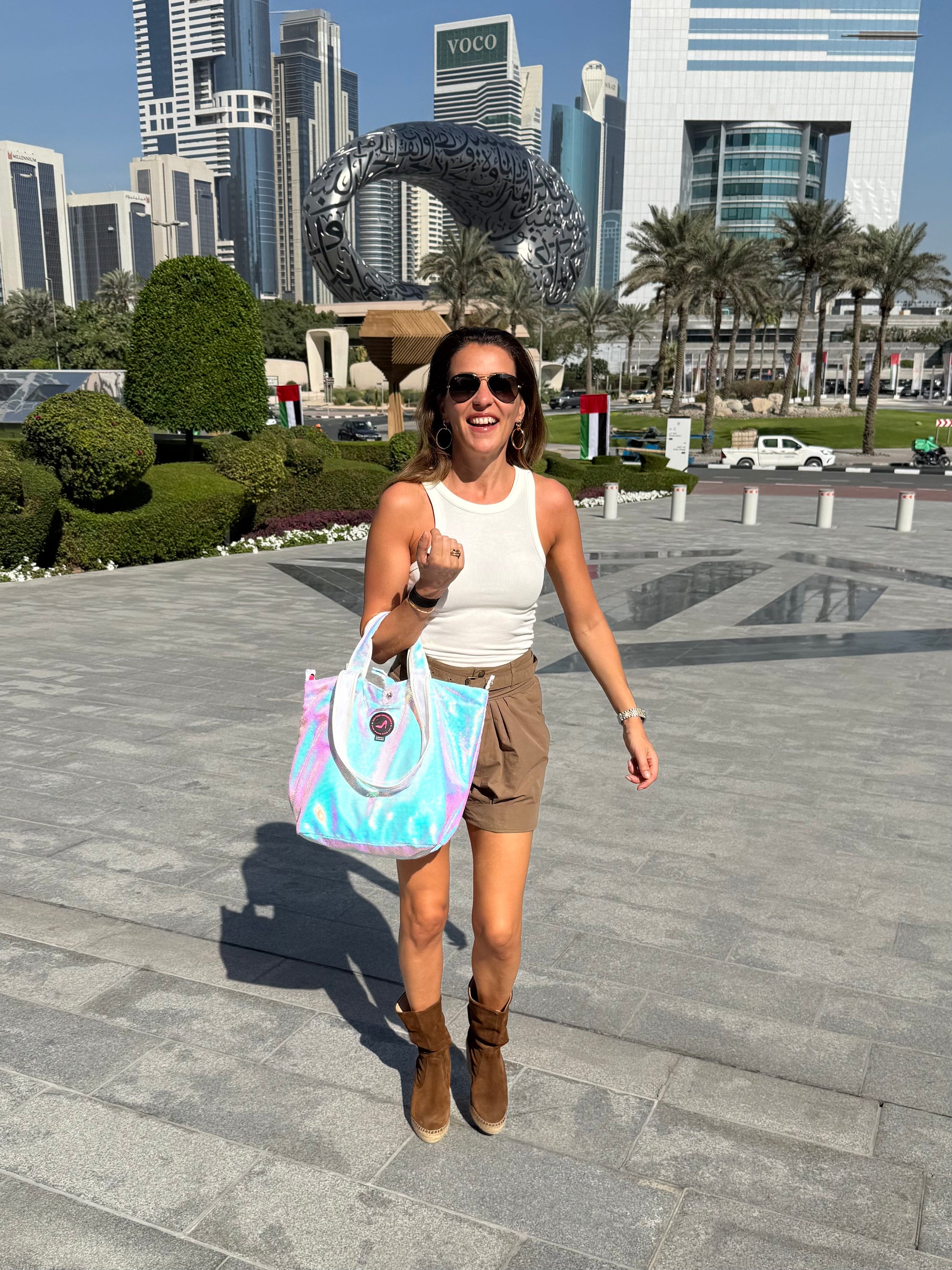 Woman walking in a Dubai city square holding an iridescent tote bag, with modern buildings and palm trees in the background
