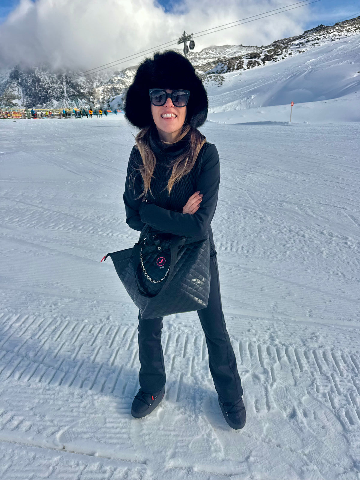 Woman in black winterwear and hat on snow carries black caviar leather small tote bag by KATE.