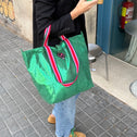 A person wearing a black blazer, blue jeans, and tan shoes holds the Small Tote Bag Metallic Green VENEZIA with striped handles, plus a phone and coffee cup, while standing on a city sidewalk near a metal chair.