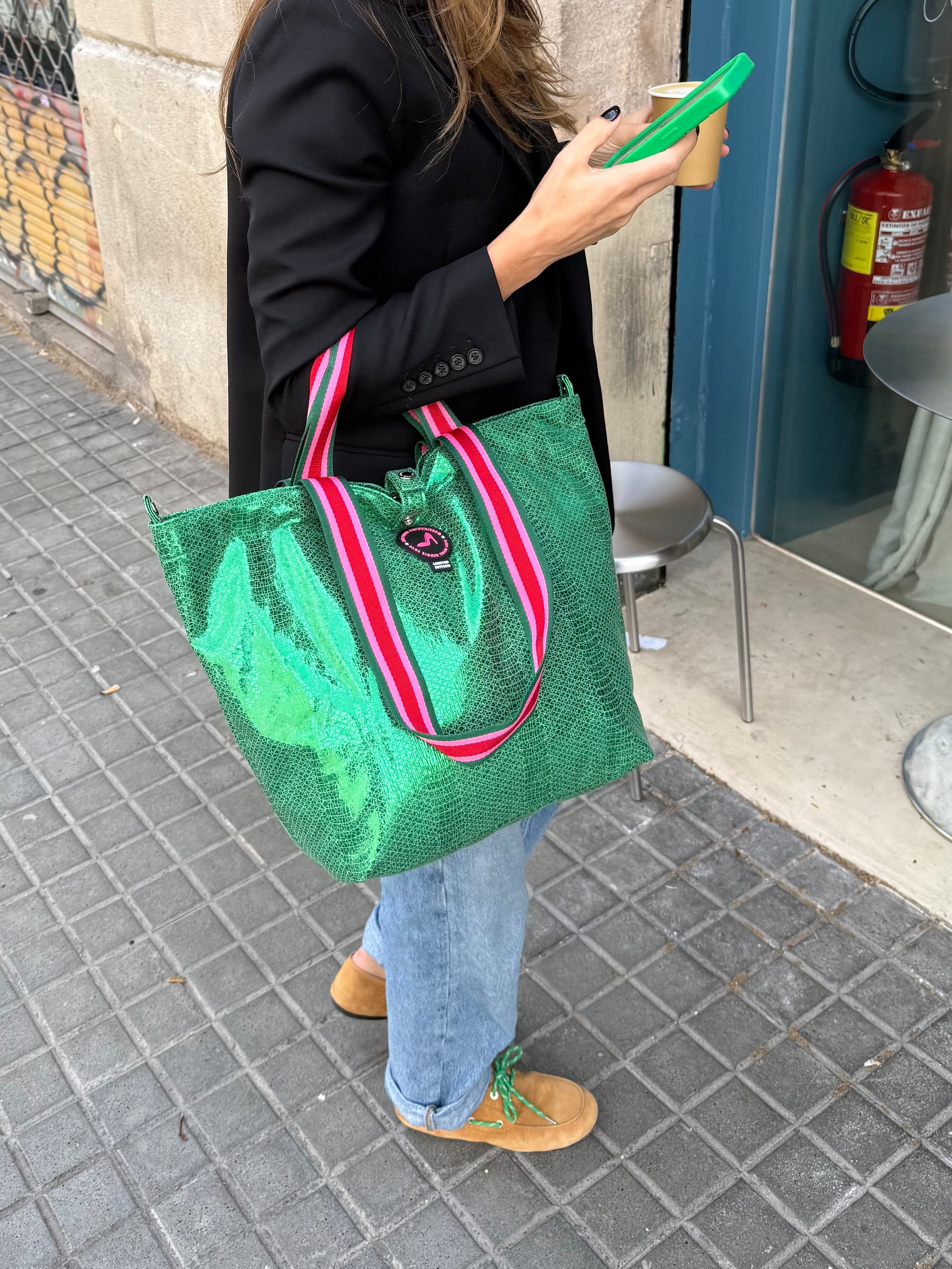 A person wearing a black blazer, blue jeans, and tan shoes holds the Small Tote Bag Metallic Green VENEZIA with striped handles, plus a phone and coffee cup, while standing on a city sidewalk near a metal chair.