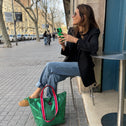 A woman at a café table smiles, holding a green phone. Next to her is the Small Tote Bag Metallic Green VENEZIA, featuring striped handles. She wears a black blazer, jeans, and loafers on a tree-lined street.