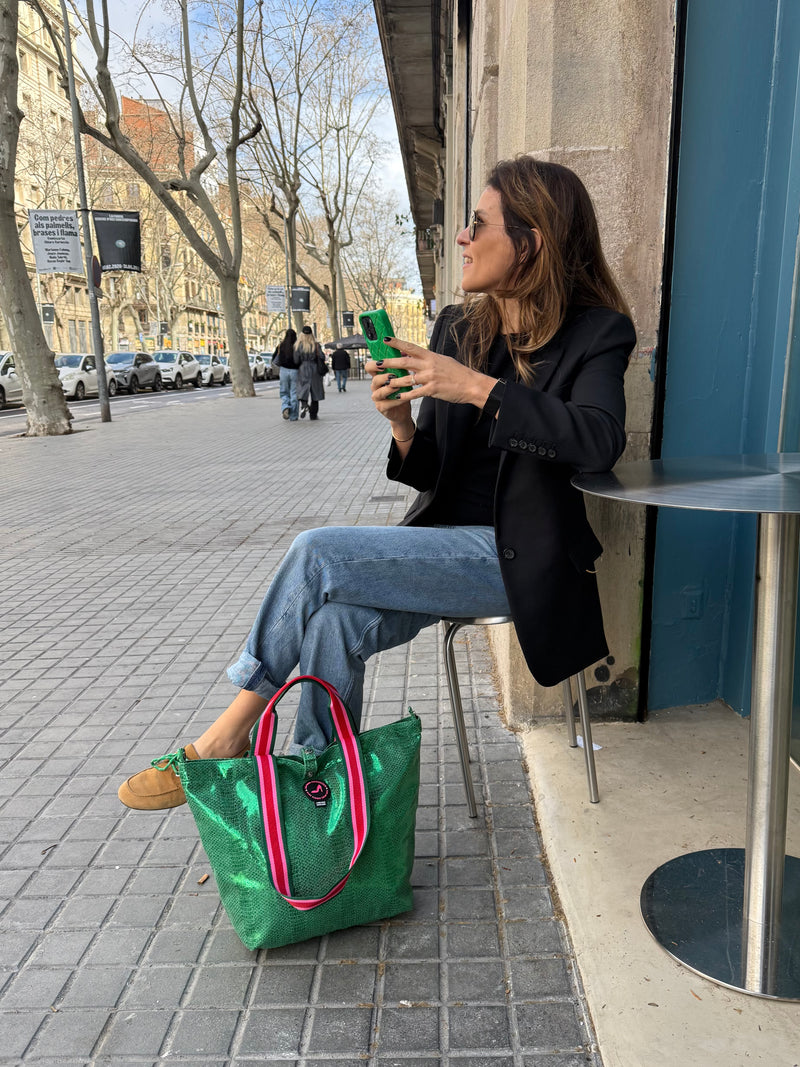A woman at a café table smiles, holding a green phone. Next to her is the Small Tote Bag Metallic Green VENEZIA, featuring striped handles. She wears a black blazer, jeans, and loafers on a tree-lined street.