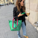 A woman stands on a city sidewalk, smiling with a coffee cup, green phone, and Small Tote Bag Metallic Green VENEZIA featuring red-and-white handles. She wears sunglasses, a black blazer, jeans, and tan shoes.