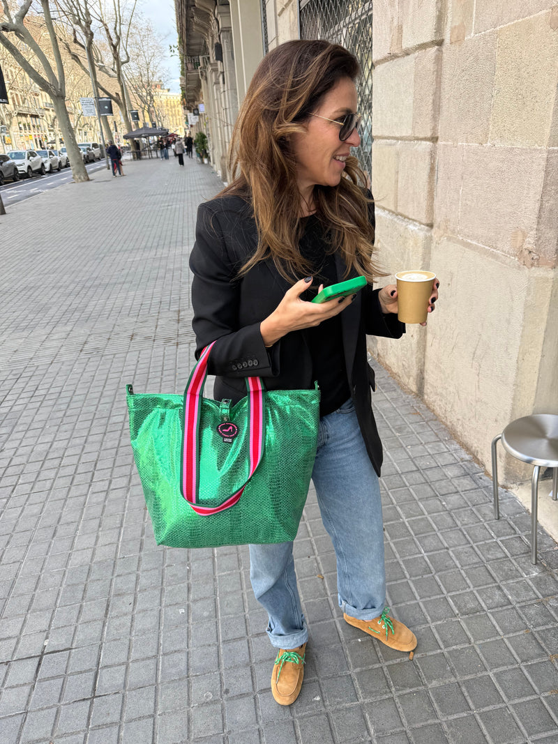 A woman stands on a city sidewalk, smiling with a coffee cup, green phone, and Small Tote Bag Metallic Green VENEZIA featuring red-and-white handles. She wears sunglasses, a black blazer, jeans, and tan shoes.