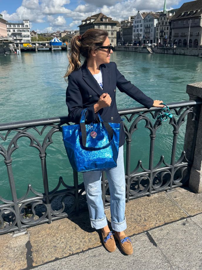 Woman holding a blue bag by a waterfront with buildings in the background