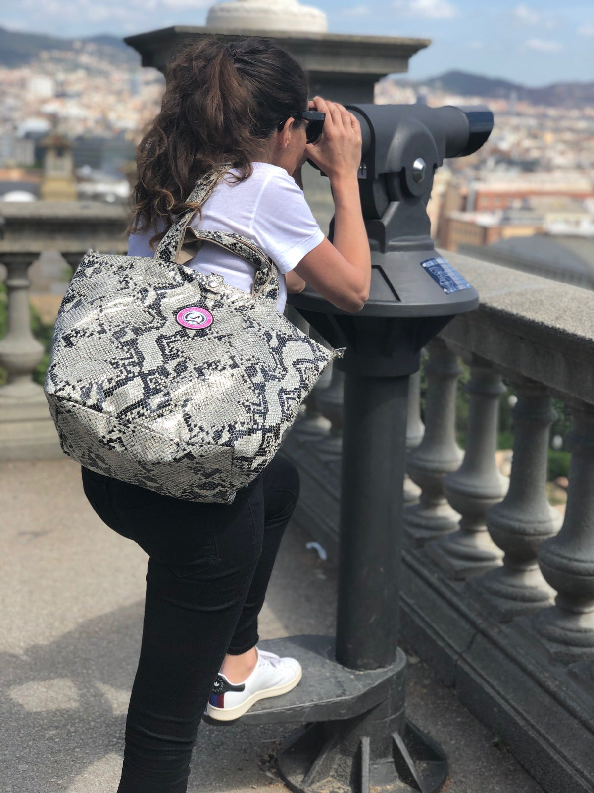 Een vrouw met een kleine draagtas met pythonprint staat bij een stadsgezichtskijker, gezien vanuit de lucht.