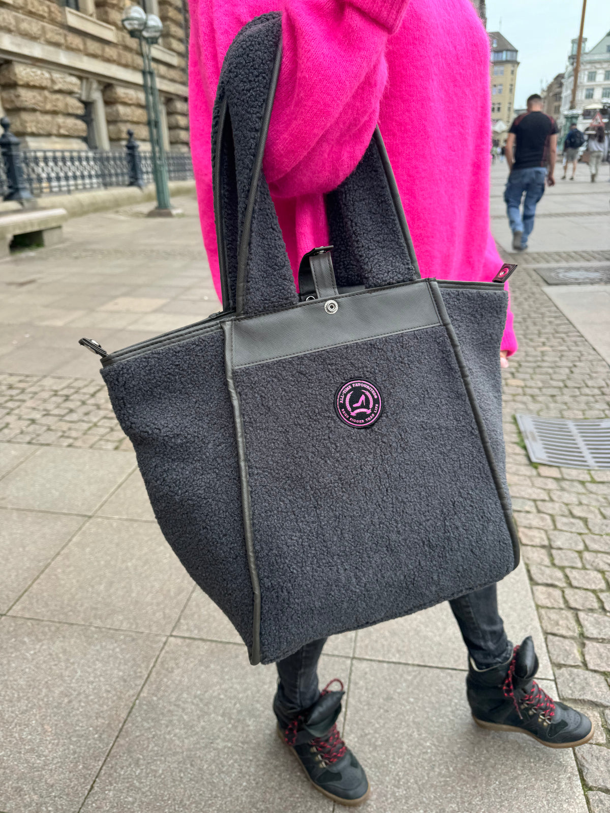 Een vrouw in een roze trui draagt een grijze ‘Teddy All-time Favourites’-draagtas met een roze patch en een ritssluiting in een stedelijke straat.