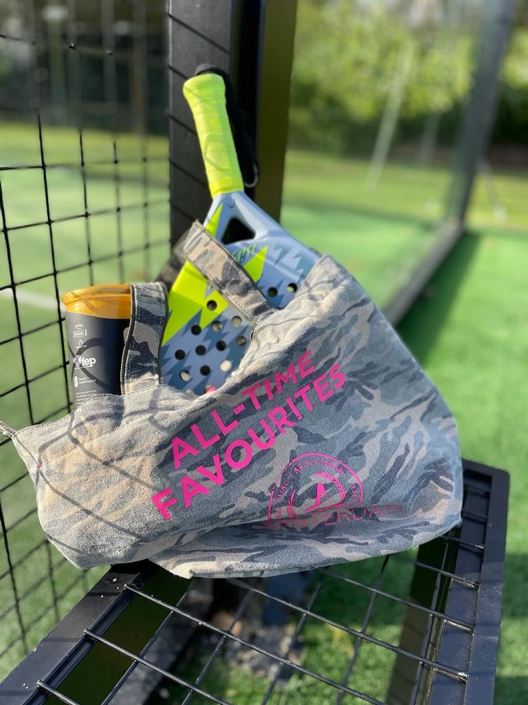 Een canvas draagtas met camouflageprint van All-time Favourites op een bankje, met daarin een fles en een peddel, vlakbij een sportveld.
