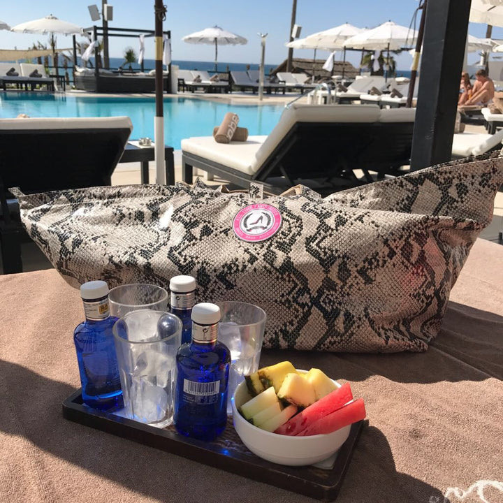 Leder-Tragetasche mit Python-Print auf einer Sonnenliege neben blauen Flaschen, Gläsern und einer Obstschale am Pool im Sonnenlicht.
