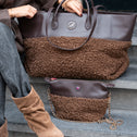 A person seated on stone steps holds a large brown tote bag with a smooth leather top and fuzzy brown lower section. A smaller matching bag with a chain strap rests on the steps below.