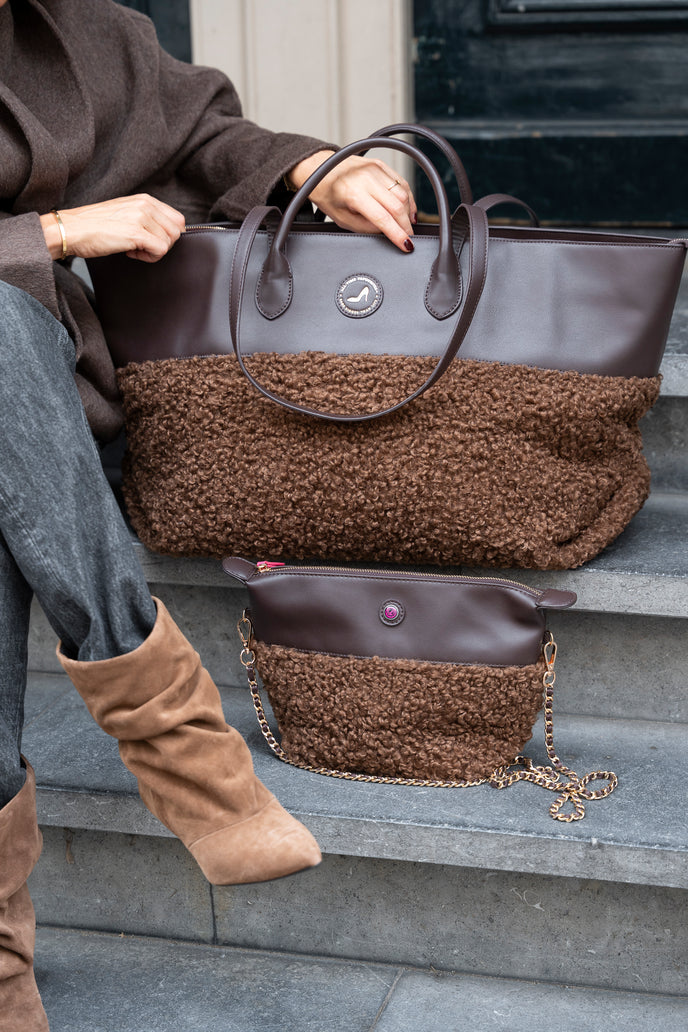 A person seated on stone steps holds a large brown tote bag with a smooth leather top and fuzzy brown lower section. A smaller matching bag with a chain strap rests on the steps below.