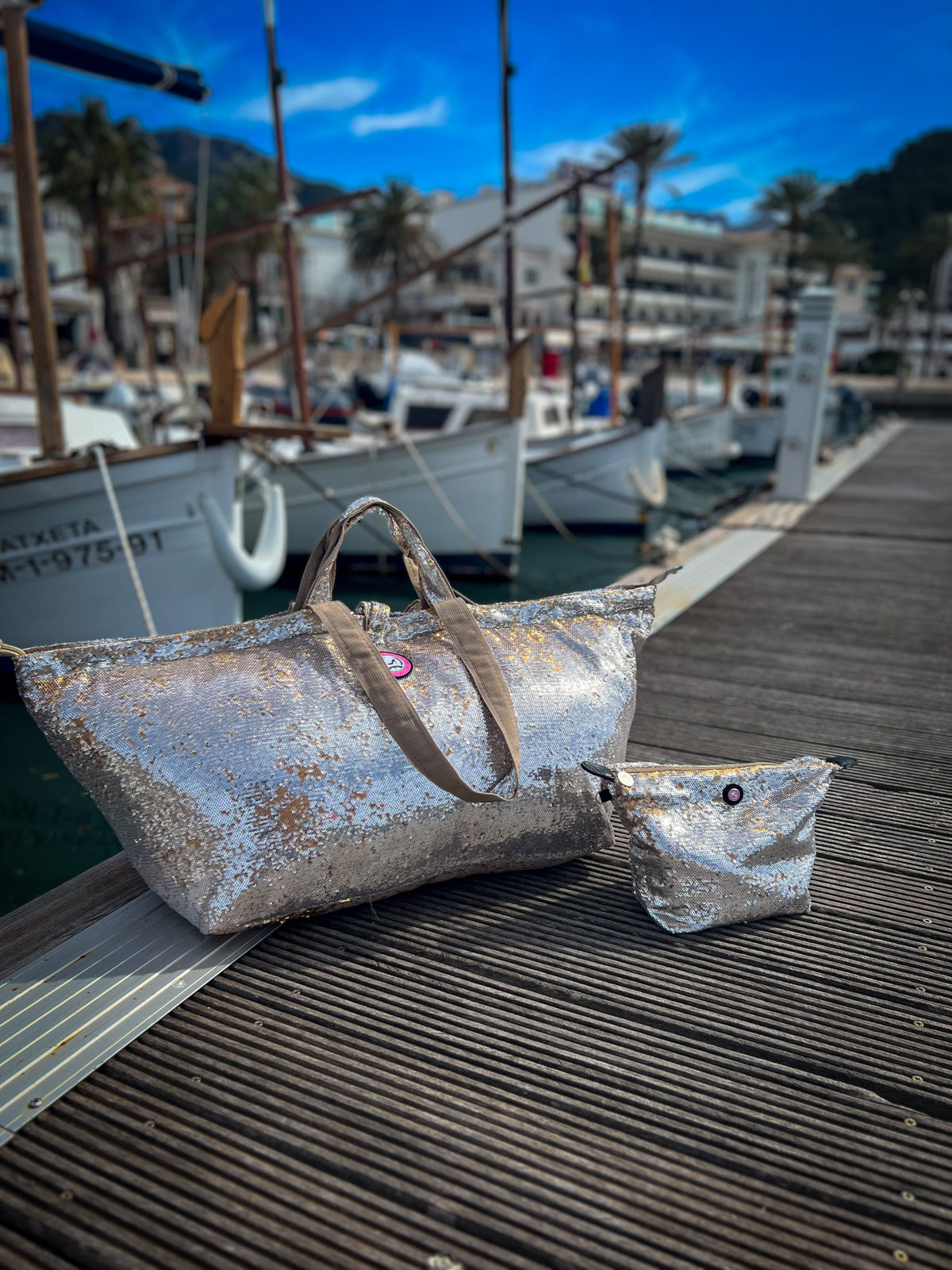 Grote Mallorca-draagtassen met zilveren en gouden pailletten van All-time Favourites op een steiger met boten en palmbomen op de achtergrond.