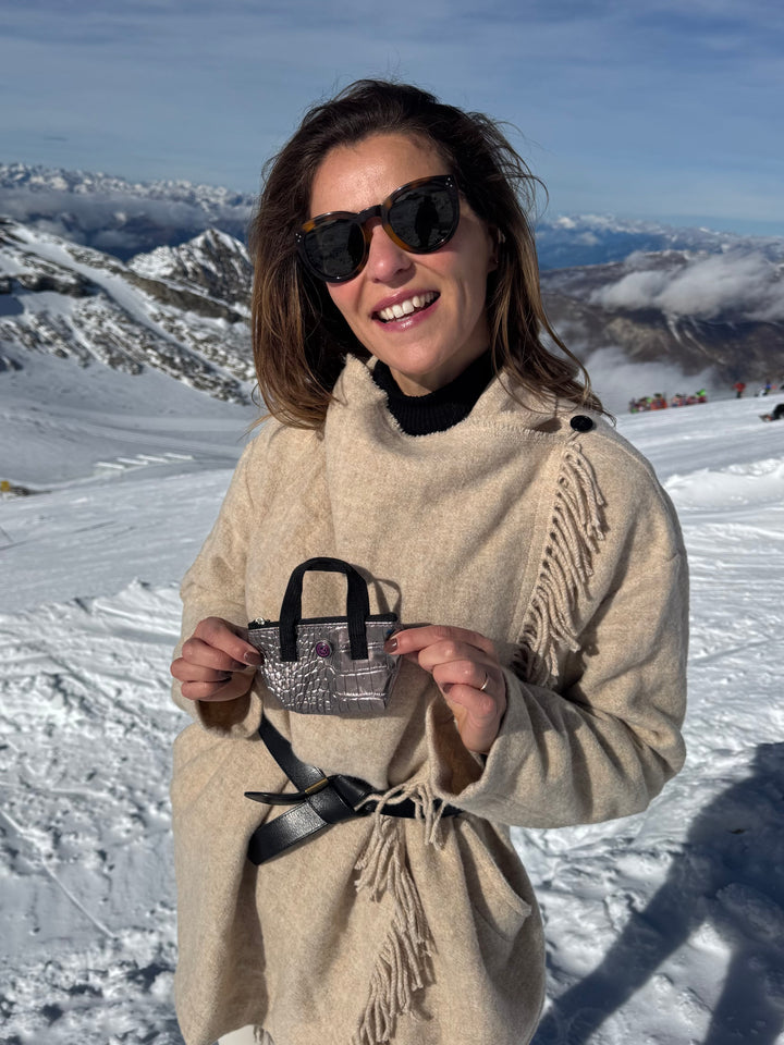 Woman in sunglasses and beige coat holding bronze metallic leather BRIGITTE key pouch on snowy mountain.
