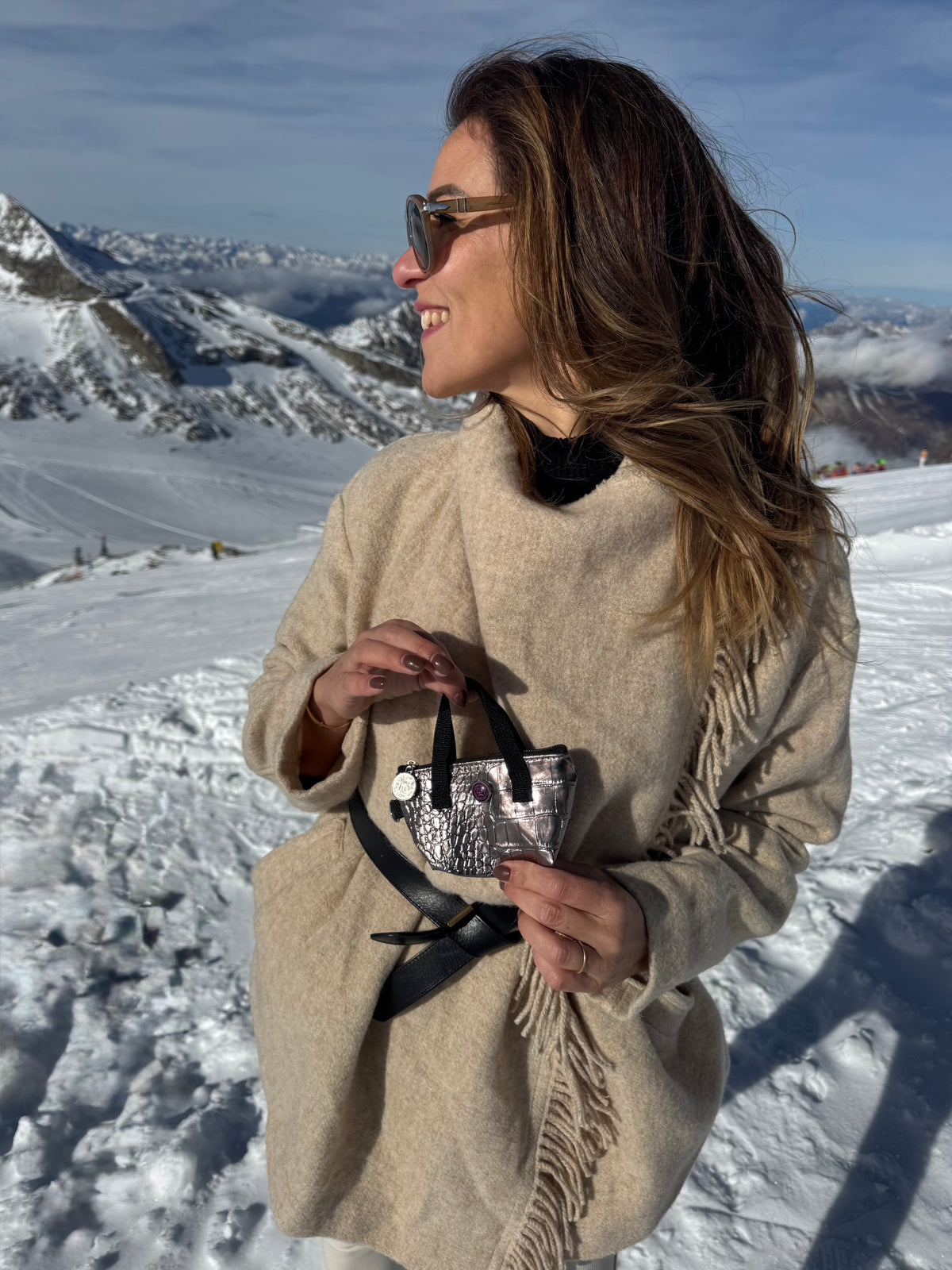 Een vrouw met een zonnebril en een beige jas houdt een sleuteltasje van bronskleurig metallic leer vast in een besneeuwd skioord.
