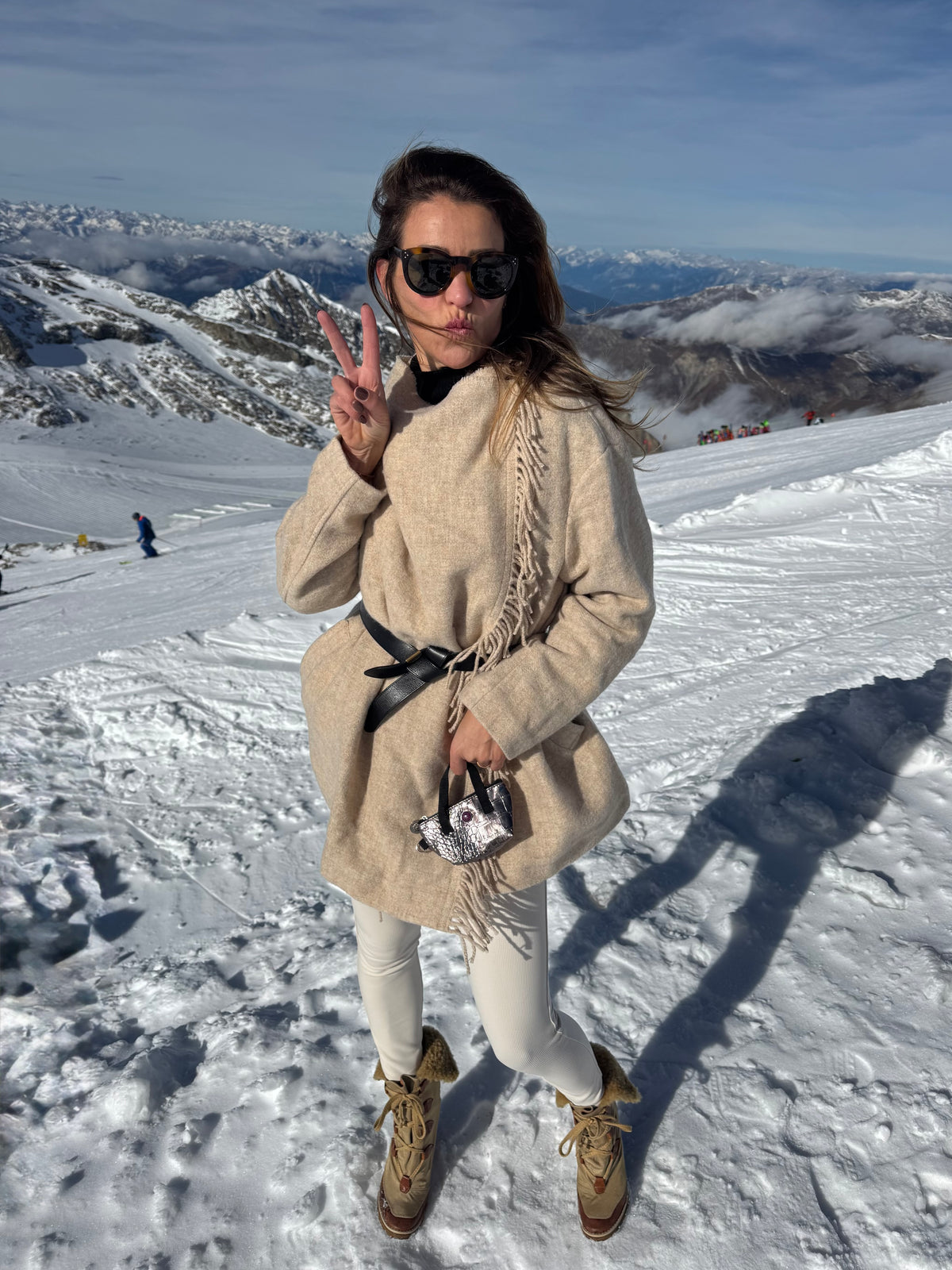 Een vrouw in een beige jas en laarzen poseert op een besneeuwde berg met het BRIGITTE-sleuteltasje van bronskleurig metallic leer.