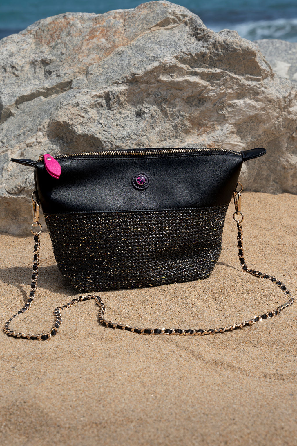Black and woven handbag on sand with rock and water background