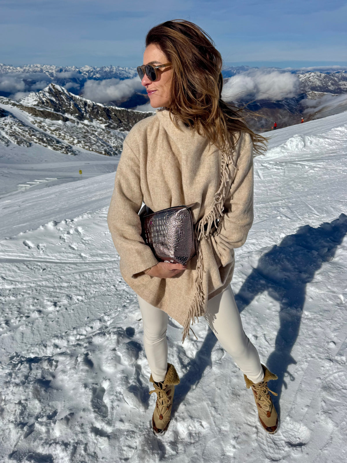 Eine Frau in einem beigen Mantel hält auf einem verschneiten Berg eine bronzefarbene, metallisch glänzende BRIGITTE-Kosmetiktasche in der Hand und trägt Stiefel.
