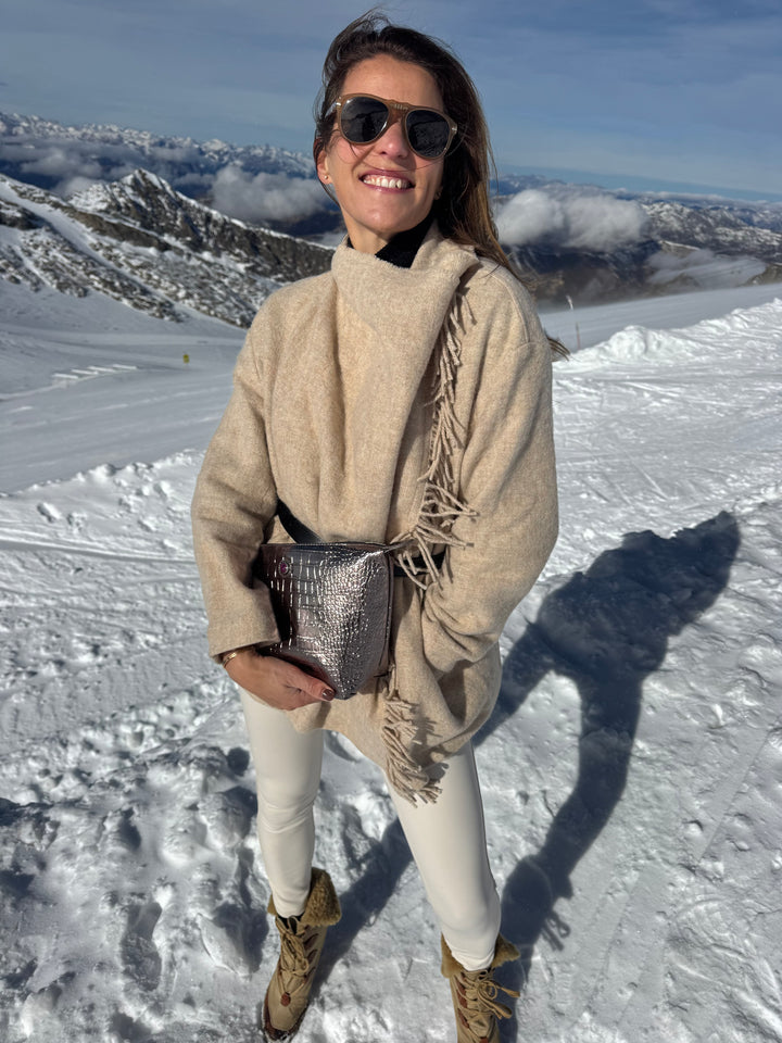 Woman in winter attire holds bronze metallic BRIGITTE toiletry bag at ski resort.