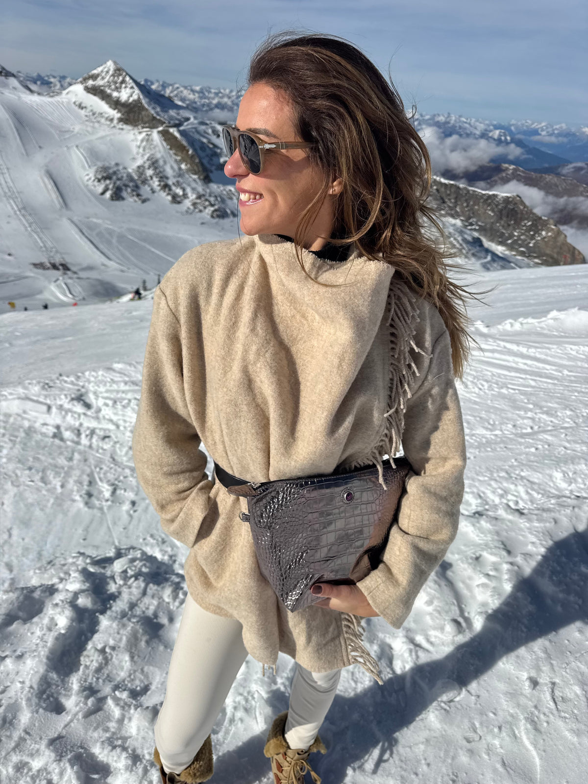 Eine Frau in einem beigen Mantel und mit Sonnenbrille auf einem schneebedeckten Gipfel, die eine BRIGITTE-Kulturbeutel aus bronzefarbenem Leder in der Hand hält.