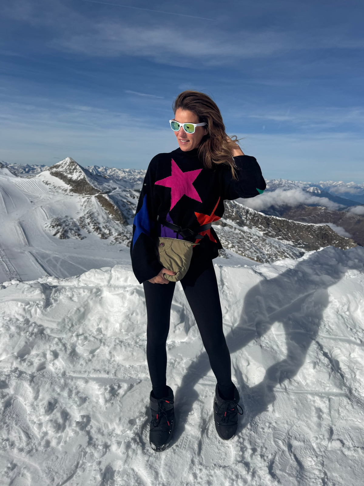 Eine Frau mit einer grünen, bauschigen Nylon-Kulturbeutel steht auf einem verschneiten Berg; sie trägt eine Sonnenbrille und einen Pullover mit Sternenmuster.