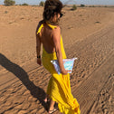 Woman in a yellow dress walking in a desert landscape holding a mermaid coloured toiletry bag