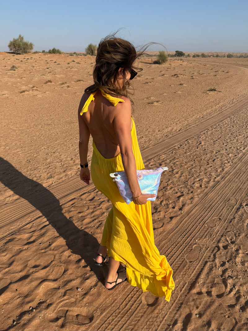 Woman in a yellow dress walking in a desert landscape holding a mermaid coloured toiletry bag