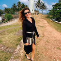 A woman in sunglasses, a black dress, and flip-flops walks near a tall white lighthouse with palm trees and blue sky, smiling as she holds the Toiletry Bag Black & White GALLE.