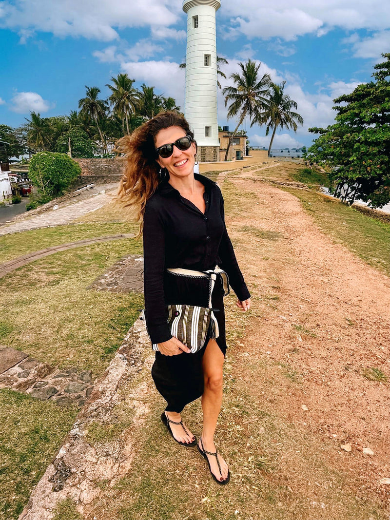 A woman in sunglasses, a black dress, and flip-flops walks near a tall white lighthouse with palm trees and blue sky, smiling as she holds the Toiletry Bag Black & White GALLE.