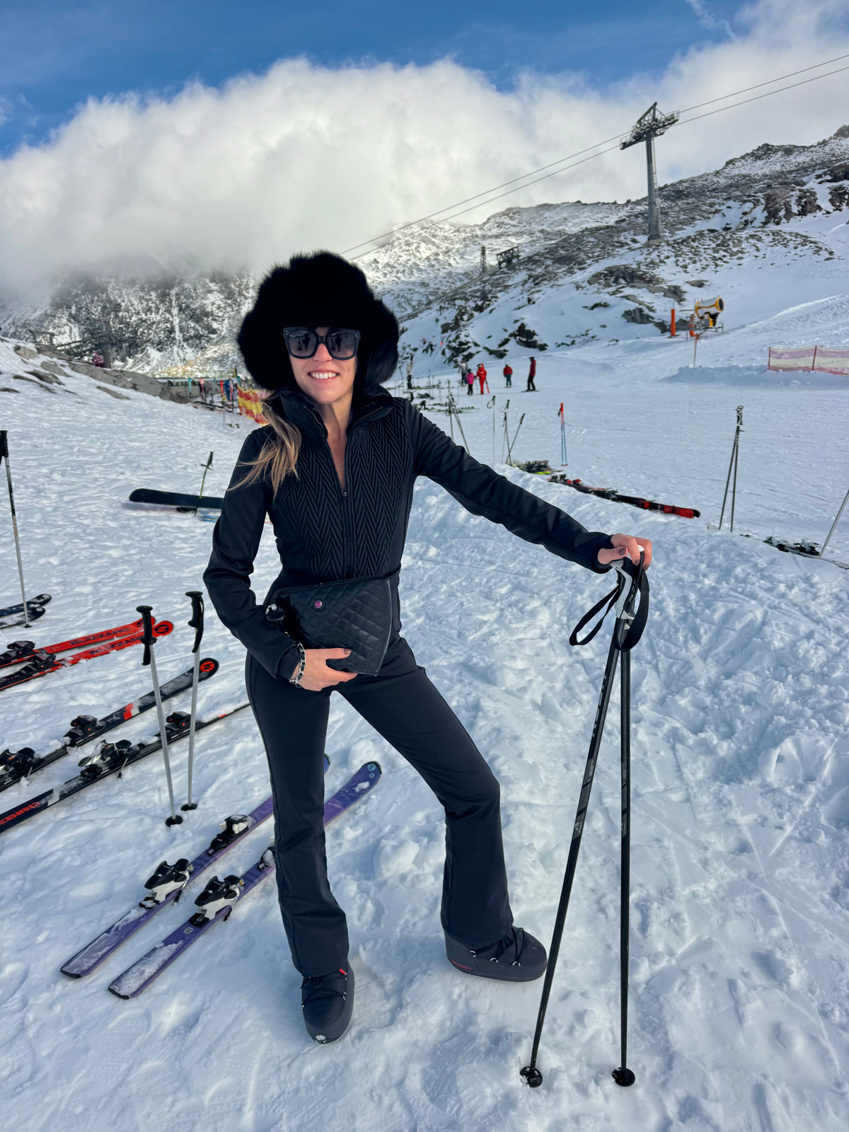 Eine Frau in schwarzer Skikleidung hält eine schwarze KATE-Kulturbeutel in der Farbe „Caviar“ in der Hand, im Hintergrund sind Skier und schneebedeckte Berge zu sehen.
