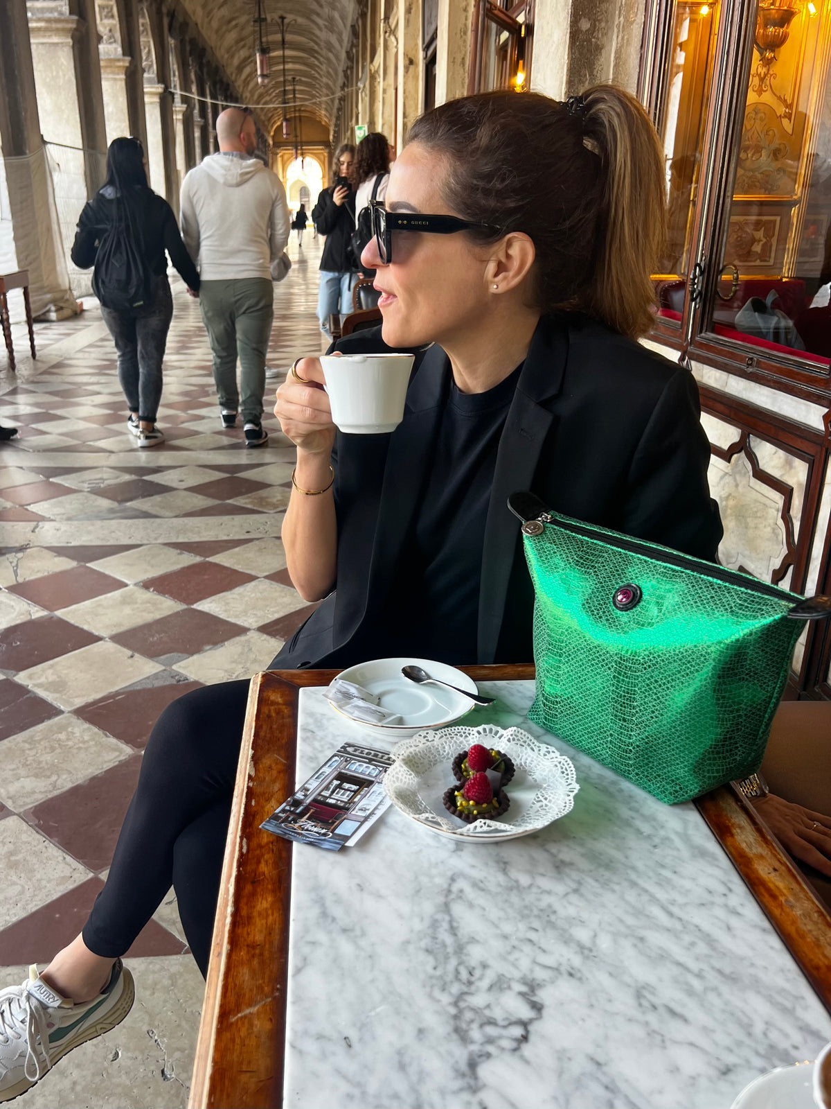 Metalen groene toilettas naast een vrouw in het zwart die buiten koffie drinkt, met een dessert op tafel, van het merk VENEZIA.