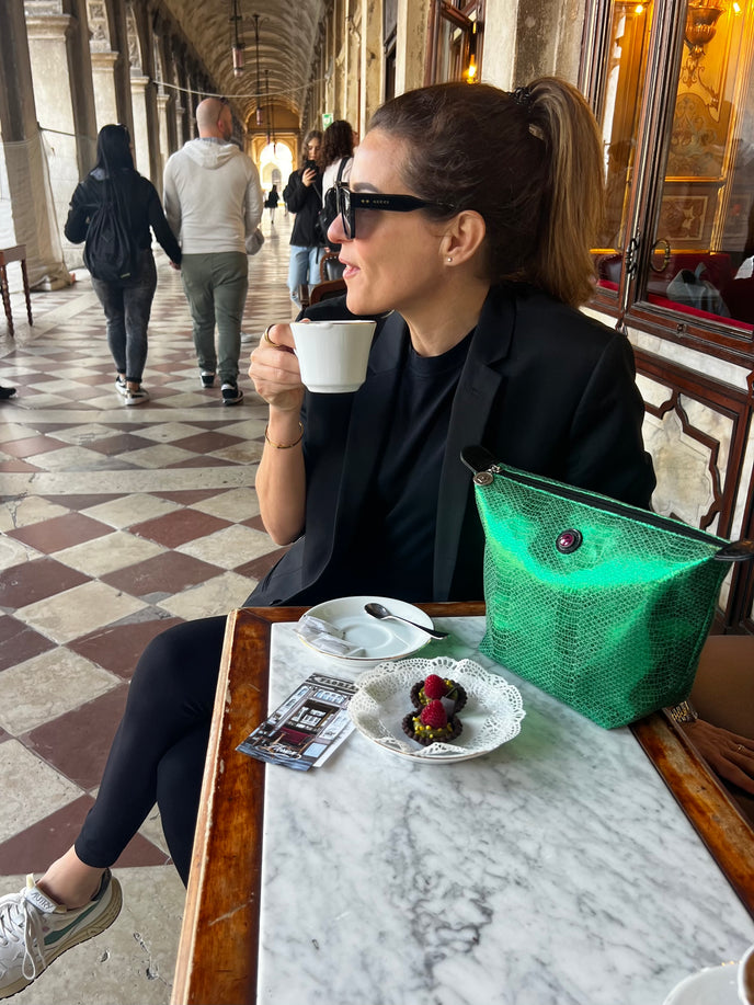 Woman enjoying coffee at a Venetian café with the Toiletry Bag Venezia in metallic green croc print on the marble table.