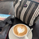 A cup of cappuccino with heart-shaped latte art rests on a saucer beside sunglasses, a book, and the stylish Toiletry Bag Black & White GALLE, all arranged on a dark wooden table.