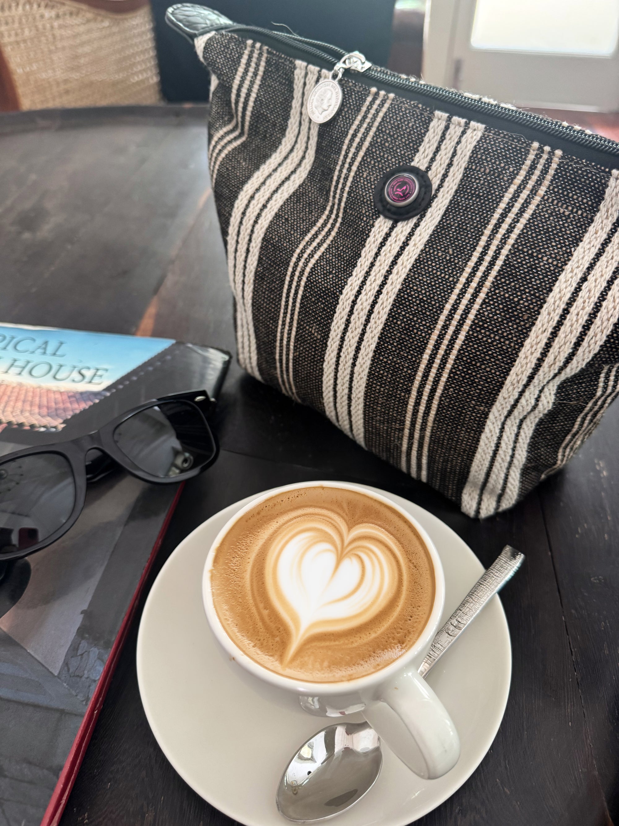A cup of cappuccino with heart-shaped latte art rests on a saucer beside sunglasses, a book, and the stylish Toiletry Bag Black & White GALLE, all arranged on a dark wooden table.