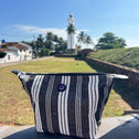 The Toiletry Bag Black & White GALLE rests on a ledge in the foreground, with a white lighthouse, palm trees, grass, and blue sky in the background.